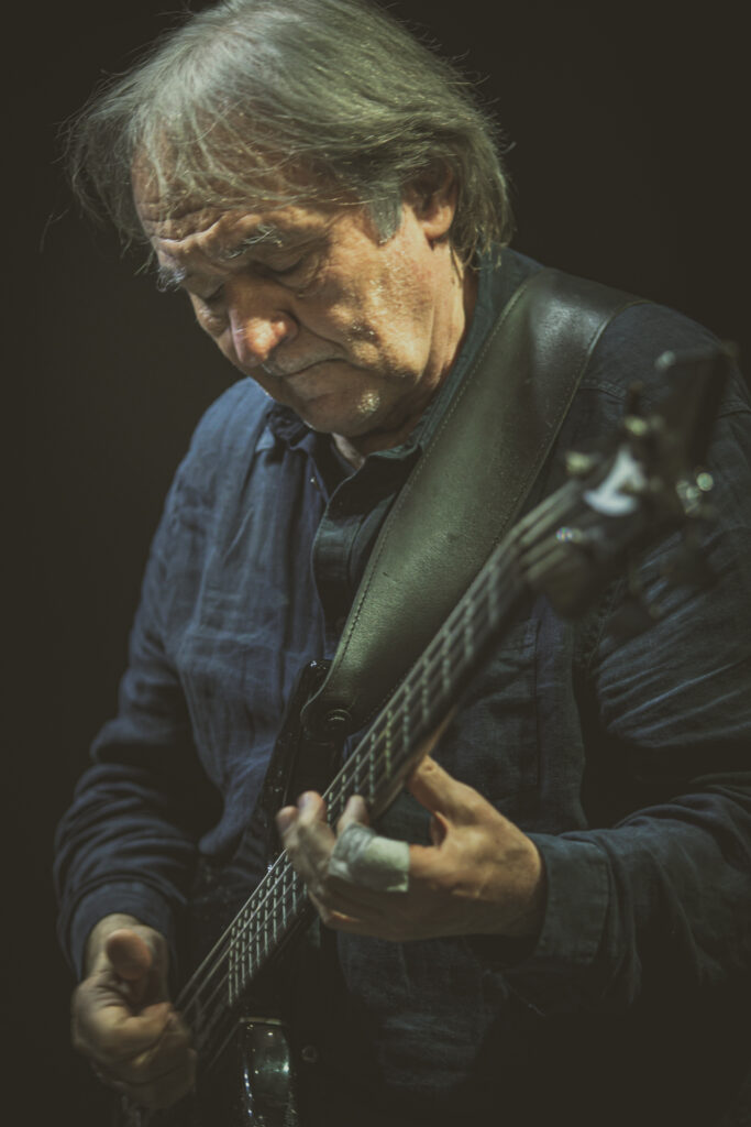Zdjęcie. Starszy mężczyzna z długimi siwiejącymi włosami gra na gitarze.