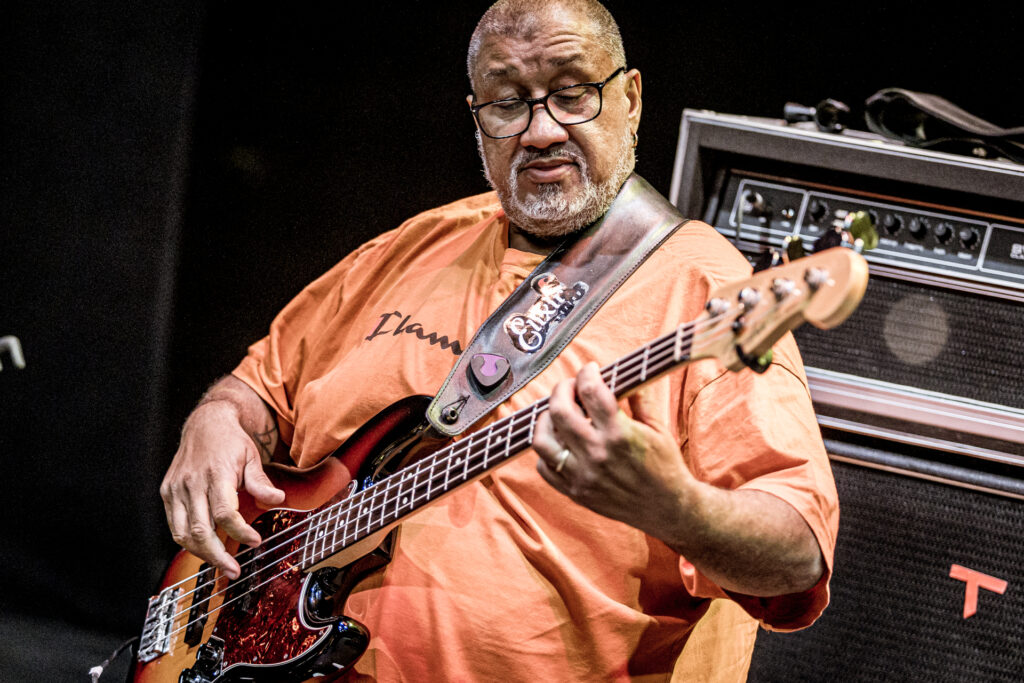 Zdjęcie. Otyły ciemnoskóry gitarzysta w pomarańczowej podkoszulce gra na gitarze.