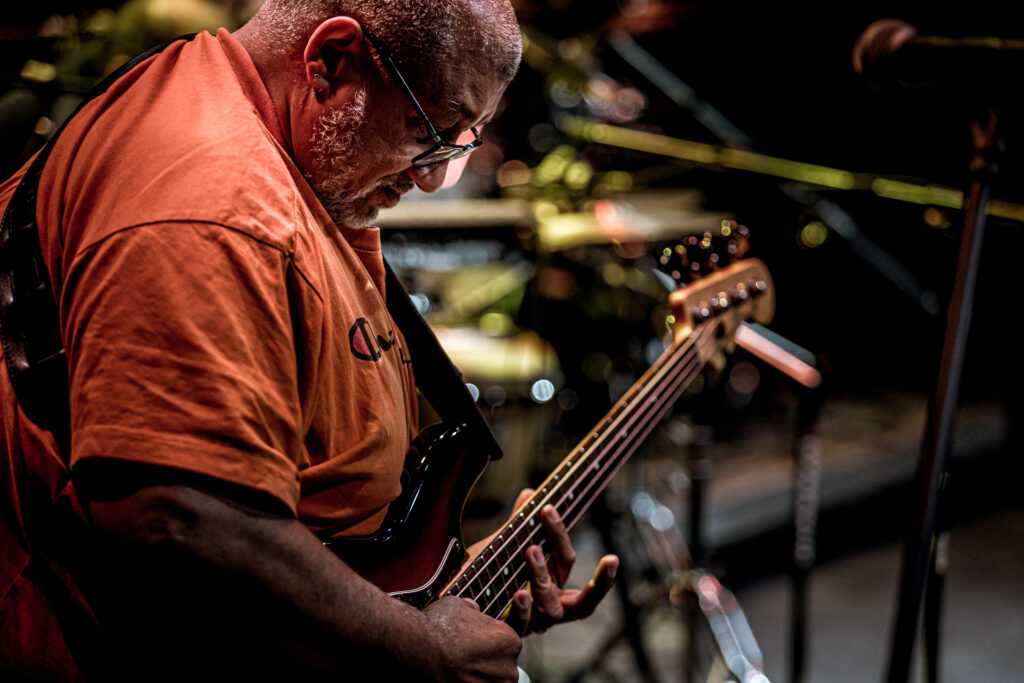 Zdjęcie. Otyły gitarzysta pochyla się nad swoim instrumentem. Gra na strunach.