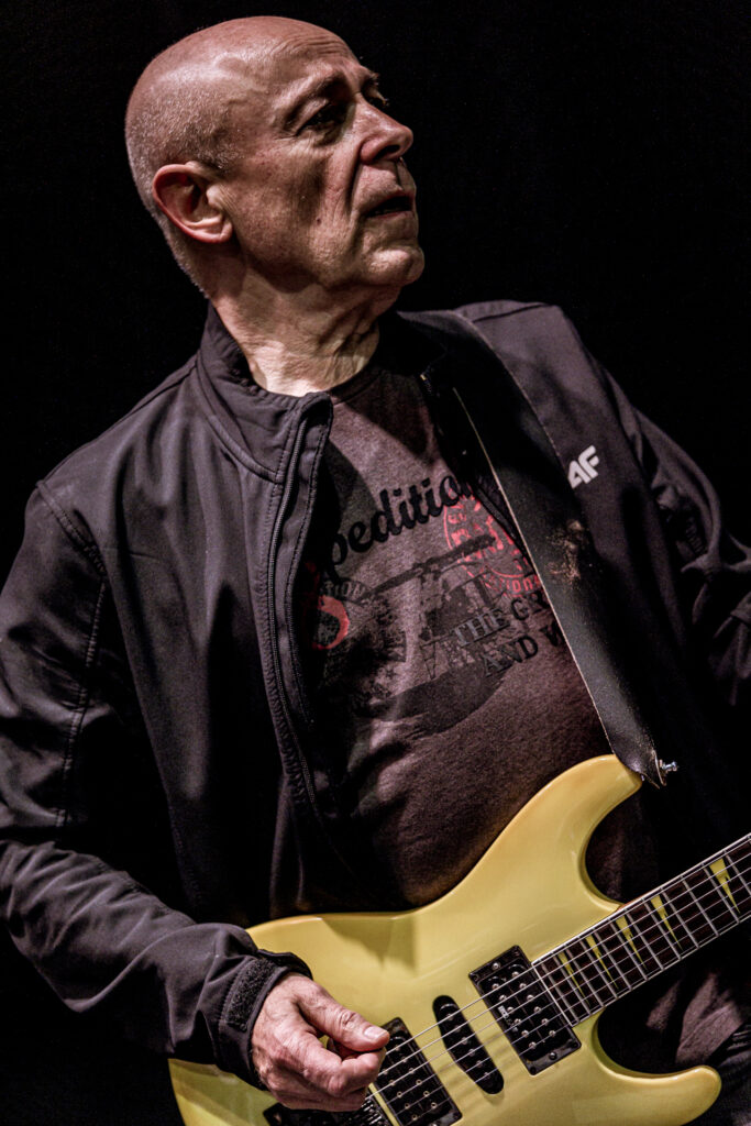 Zdjęcie. Łysy gitarzysta gra na żółtej gitarze.