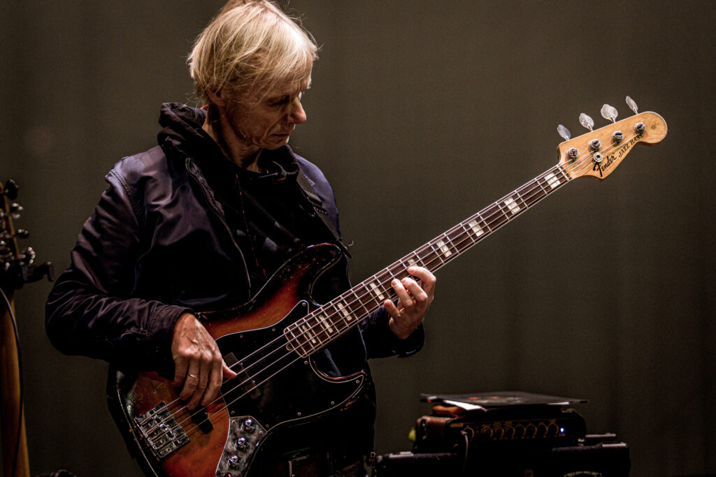Zdjęcie. Mężczyzna p włosach blond w fioletowej kurtce gra na gitarze elektrycznej.