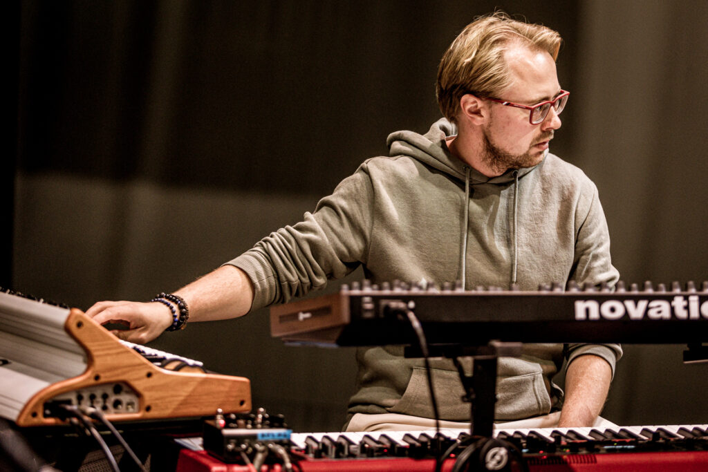 Zdjęcie. Muzyk z dłuższą blond grzywką i czerwonymi oprawkami okularów gra na keyboardzie.