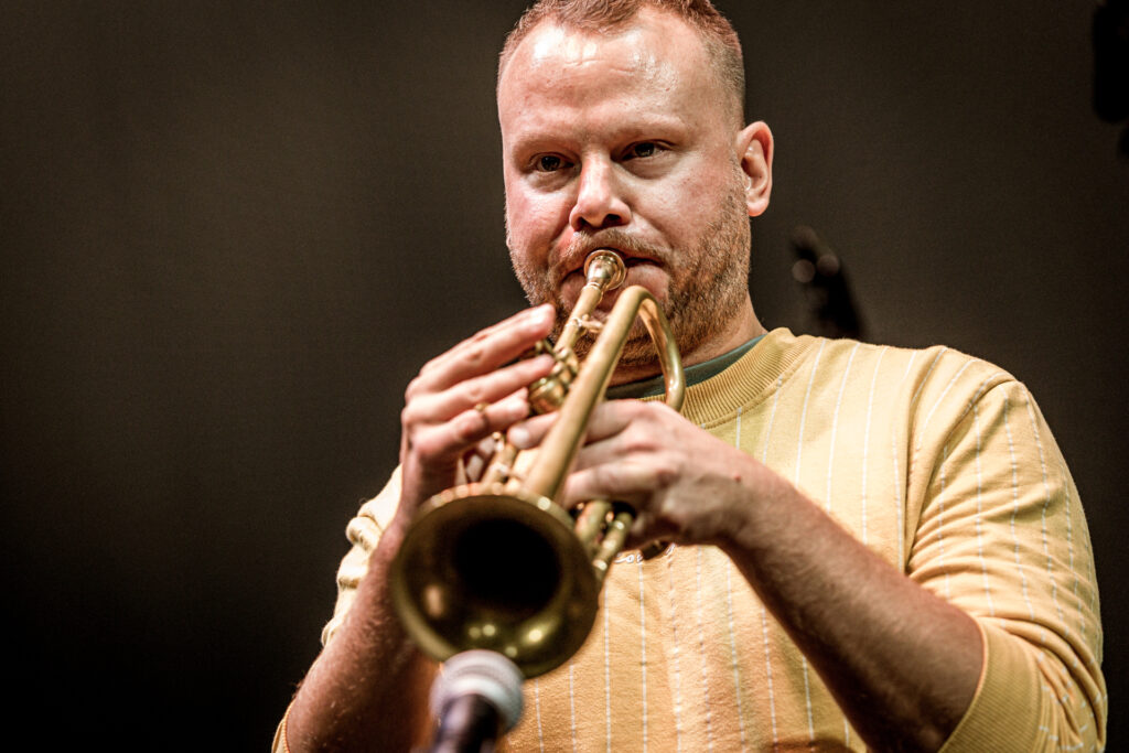 Zdjęcie. Rudowłosy trębacz w żółtej bluzie gra na instrumencie.