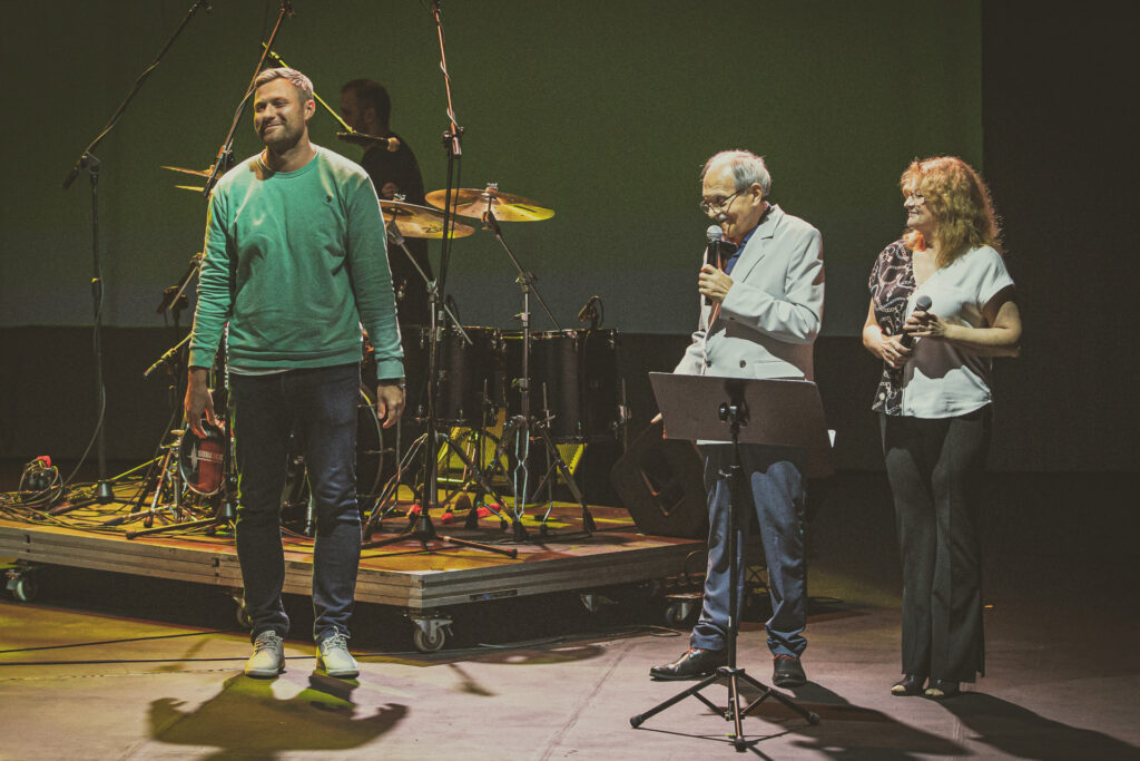 Zdjęcie. Wysoki młody uśmiechnięty mężczyzna na scenie KCK. To koordynujący festiwal Memorial to Miles Krzysztof Zięba z impresariatu KCK. Obok prowadzący festiwal Marzena i Leszek Ślusarczykowie.
