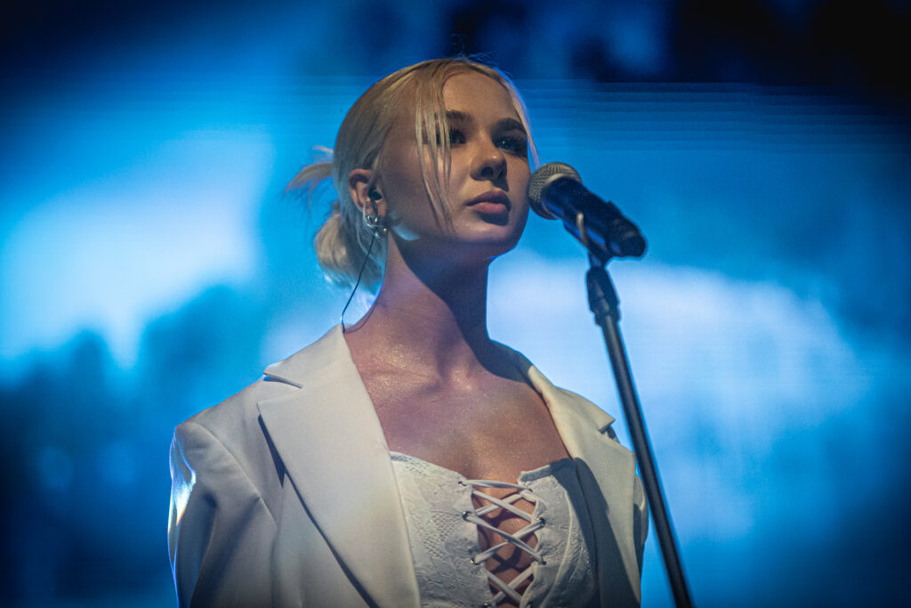 Zdjęcie. Blondynka w jasny stroju stoi przed mikrofonem.