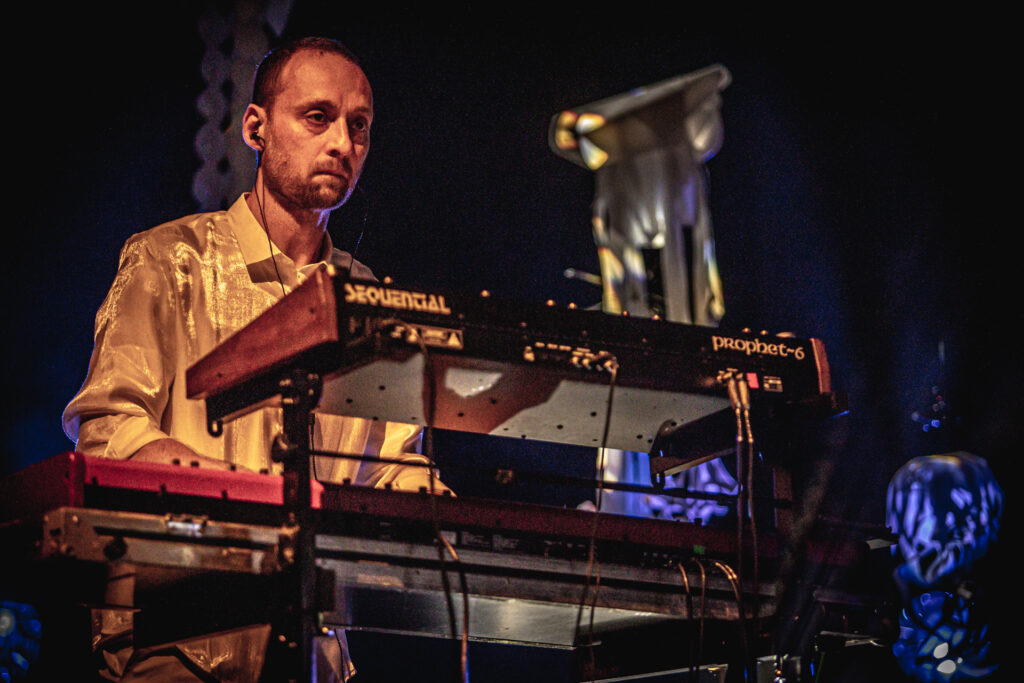 Zdjęcie. Mężczyzna z ciemną krótką brodą i wąsami gra na keyboardzie.