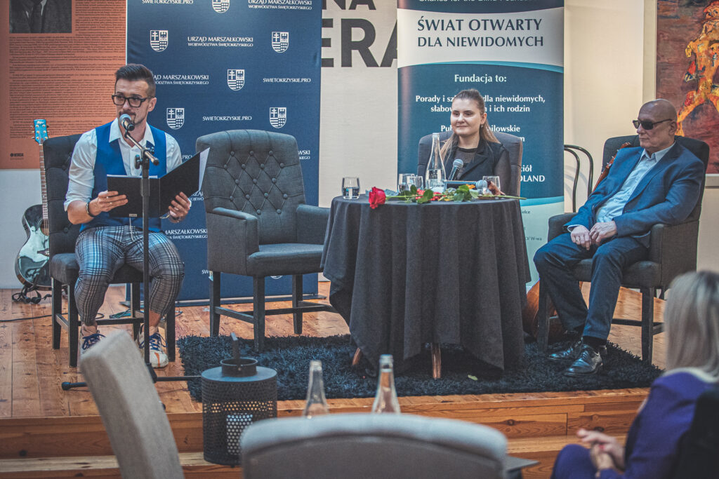 Zdjęcie. Na podwyższeniu w galerii Winda przy stole siedzą trzy osoby. Jedna kobieta i dwoóh mężczyzn. Jeden młody i szczupły czyta do mikrofonu. Drugi jest łysy i ma na nosie ciemne okulary.