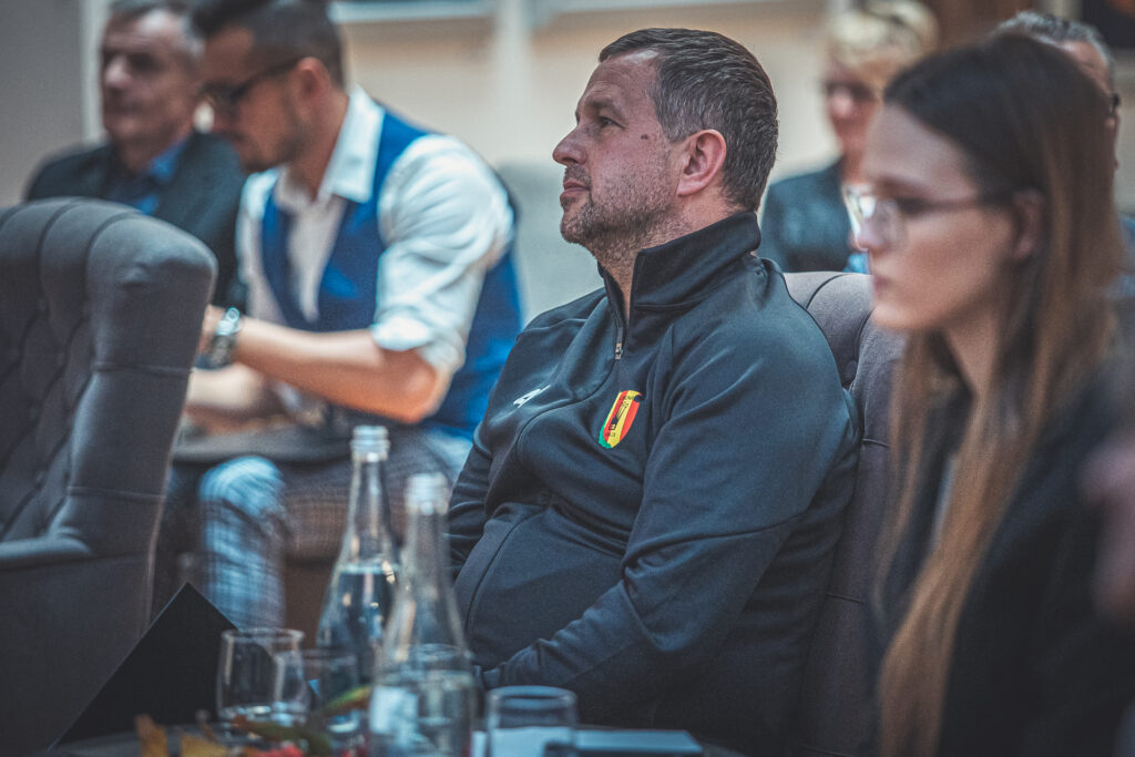 Zdjęcie. Mężczyzna w dresie z logotypem klubu Korona Kielce siedzi przy stoliku podczas spotkania z Markiem Kalbarczykiem.
