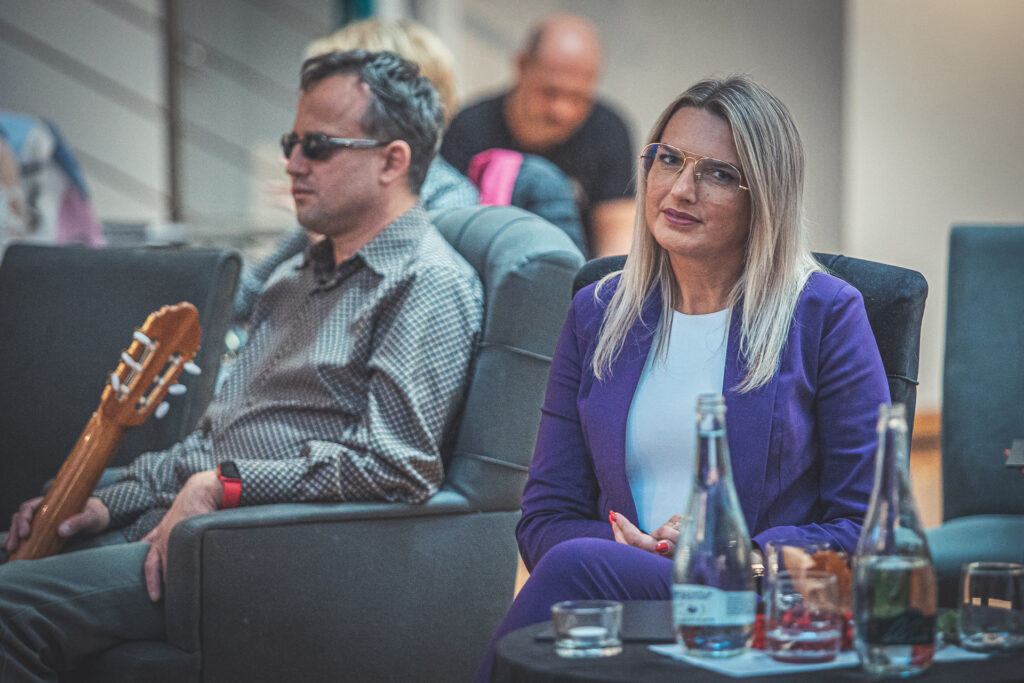 Zdjęcie. Radna Joanna Winiarska i mężczyzna z gitarą siedzą przy okrągłych stolikach w Galerii Winda.