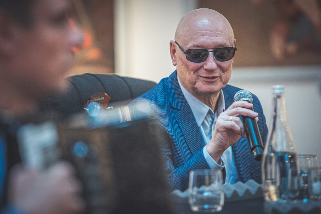 Zdjęcie. Marek Klbarczyk - łysy mężczyzna w ciemnych okularach podczas spotkania w Galerii Winda mówi do mikrofonu.