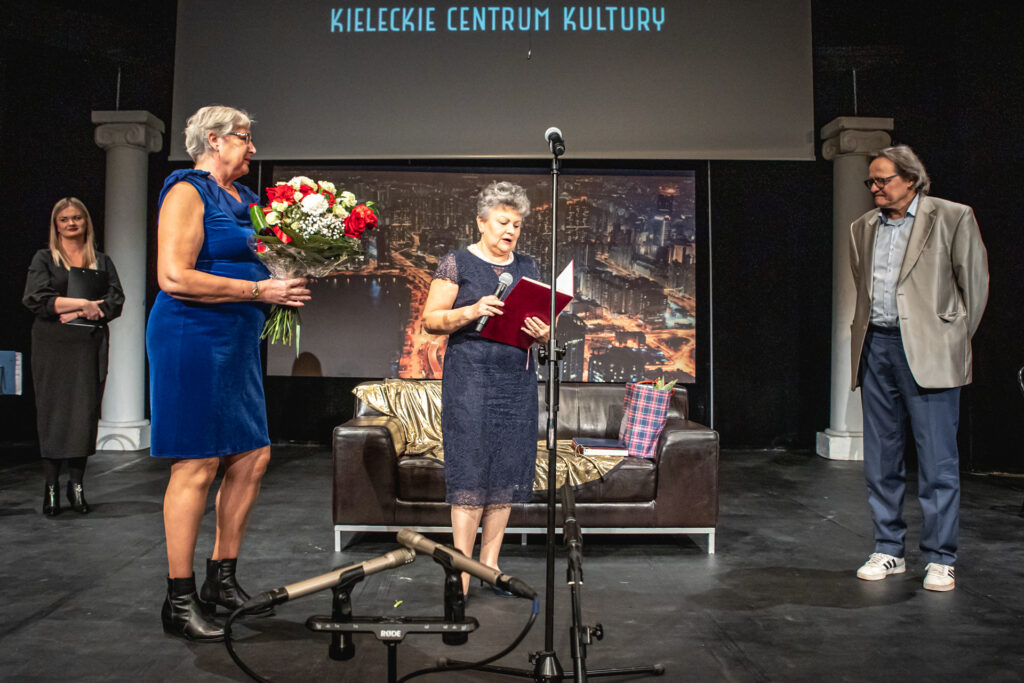 Zdjęcie. Kobieta w koronkowej granatowej sukience czyta list gratulacyjny z okazji 25-lecia Teatru Pegaz. Obok pani w średnim wieku z bukietem kwiatów. Po prawej Andrzej Skorupski.