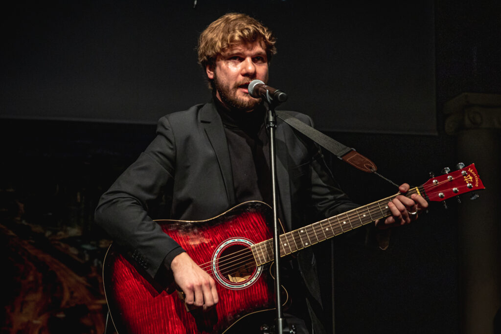 Zdjęcie. Aktor Karol Czajkowski gra na gitarze i śpiewa.