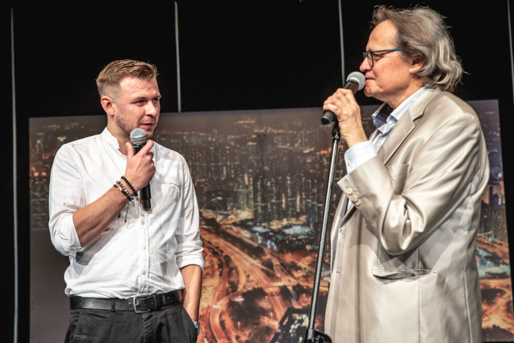 Zdjęcie. Andrezj Skorupski mowi do mikrofonu. Obok młodszy mężczyzna w białej koszuli z mikrofonem w ręku.