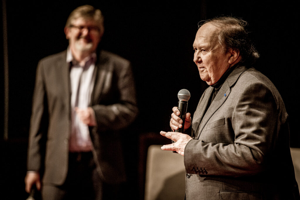 Zdjęcie. Pomysłodawca festiwalu Krzysztof Miklaszewski mówi do mikrofonu. W tle kierownik impresariatu Robert Wieczorek.