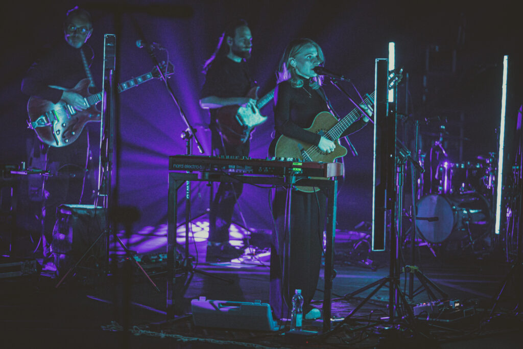 Zdjęcie. Koncert Kaśki Sochackiej. SCena tonie w pólmroku, oświetlona fioletowym światłem scenicznym. Przy mikrofonie wokalistka z gitarą. W tle dwóch muzyków.
