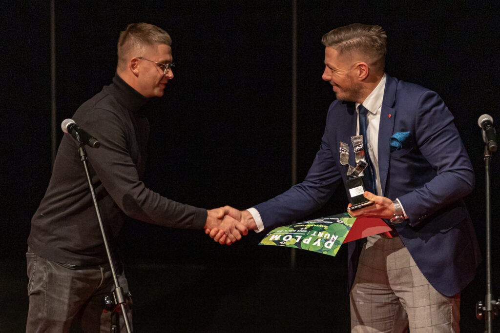 Zdjęcie. Wiceprezydent Marcin Chłodnicki z uśmiechem gratuluje nagrody mężczyźnie w czarnym golfie i okularach.