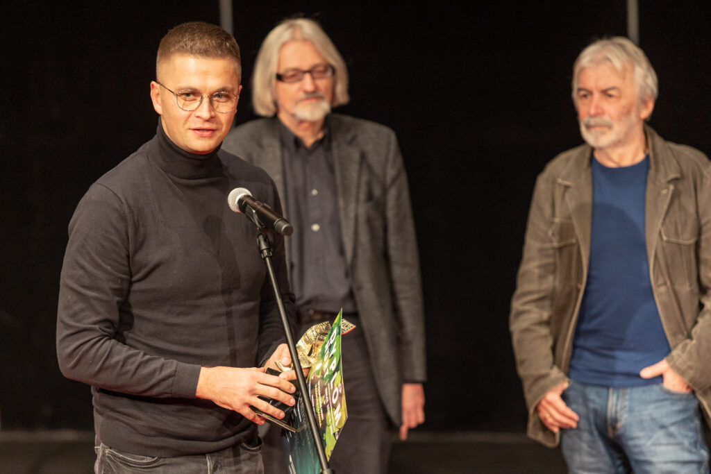 Zdjęcie. Mężczyzna w czarnym golfie mówi do mikrofonu. W tle członkowie jury festiwalu Nurt.