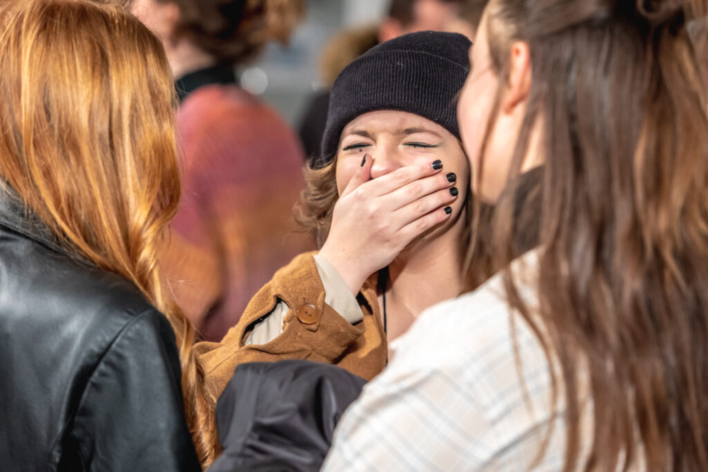 Zdjęcie. Młoda dziewczyna w czarnej czapce zaslania dłonią usta,. Przed nią dwie długowłose dziewczyny