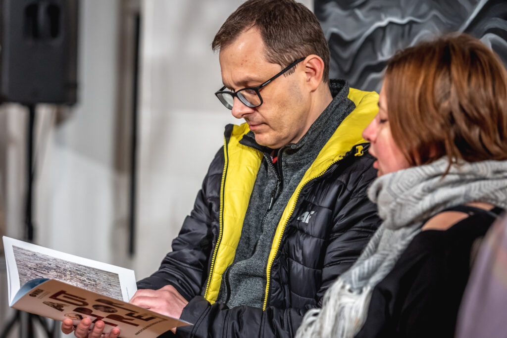 Zdjęcie. Dwójka osób: mężczyzna i kobieta stoją tyłem do jednej z prac. Mężćzyzna ogląda katalog wystawy Autoportret