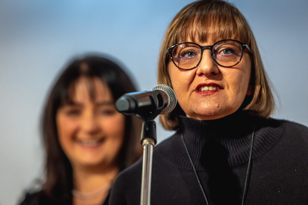 Zdjęcie. Kobieta w okularach o ciemnych oprawkach z grzywką zaslaniającą czoło i prostymi wlosami do ramion mówi do mikrofonu. To kuratorka wystawy Iwona Stachura.