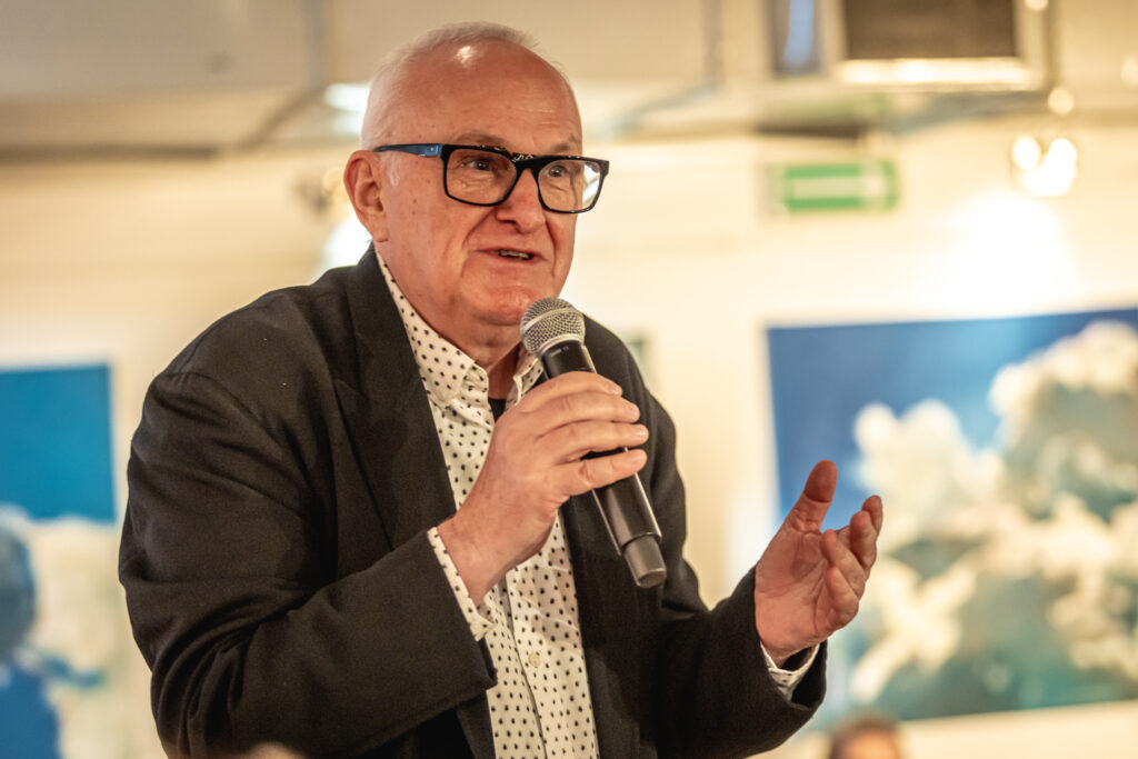 Jacek Cygan mówi do mikrofonu. Mężczyzna jest starszy, łysawy, ma okulary w ciemnych oprawkach na nosie, jasną koszulę w drobny czarny deseń i ciemną marynarkę.