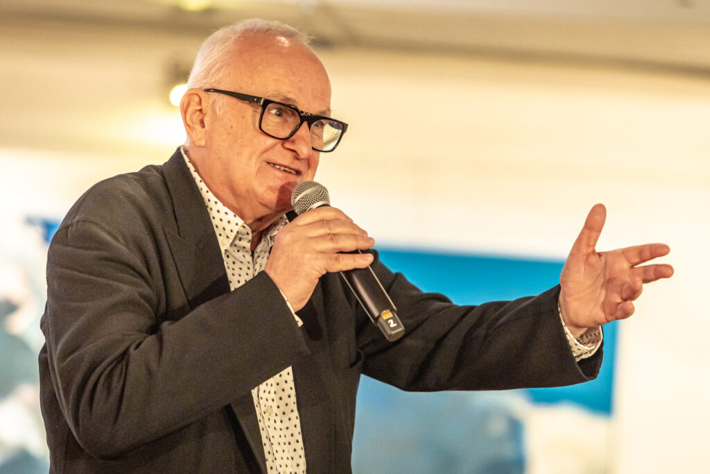 Jacek Cygan mówi do mikrofonu. Mężczyzna jest starszy, łysawy, ma okulary w ciemnych oprawkach na nosie, jasną koszulę w drobny czarny deseń i ciemną marynarkę. Lewą rękę wyciąga w stronę publiczności.