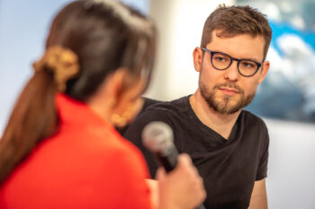 Zdjęcie. Dwie osoby rozmawiają ze sobą. Przodem do obiektywu młody mężczyzna w okularach i z brodą. To Maciej Makselon. Tyłem - kobieta w czerwonej marynarce z ługimi ciemnymi włosami. To Magdalena Fudala. Zdjęcie z "Salonu kobiet" - spotkanie z Maciejem Makselonem.