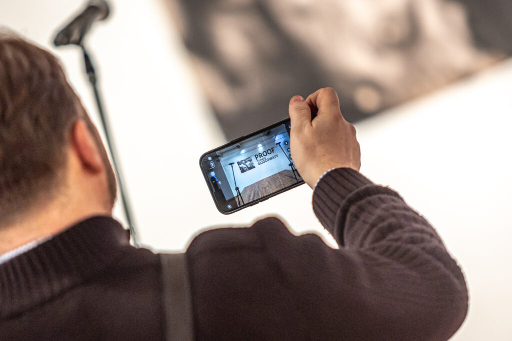 Zdjęcie. Mężczyzna fotografuje komórką ścianę z napisem Proof Tomasz Gudzowaty. Kadr można zobaczyć na wyświetlaczu jego telefonu. .