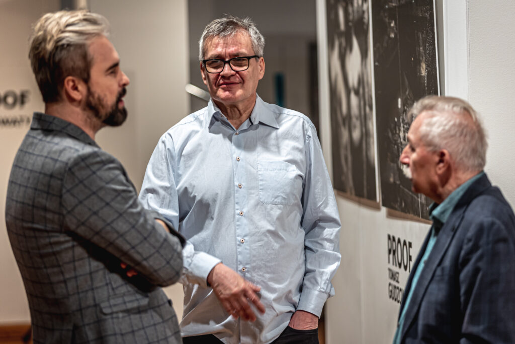 Zdjęcie. Trzech mężczyzn stoi pod ścianą, na której wiszą dż e prace Tomasza Gudzowatego. Jeden z nich to wicedyrektor Artur Wijata.
