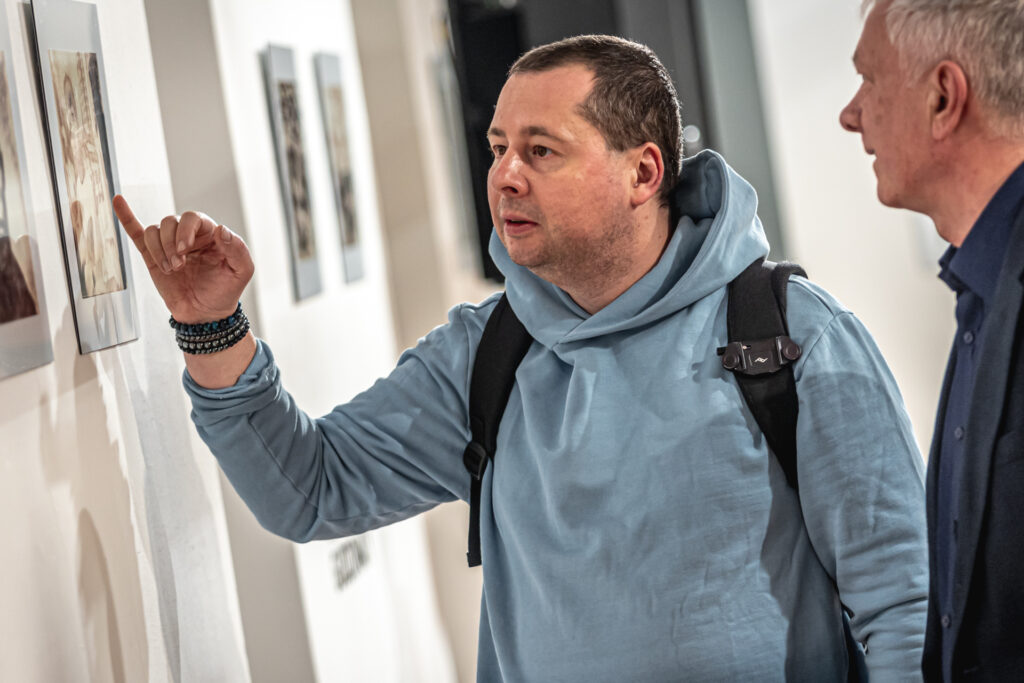 Zdjęcie. Dwaj mężczyżni przyglądają się pracom Tomasza Gudzowatego. Jeden z nich w jasnoniebieskiej bluzie wyciąga prawą dłoń i najmniejszy palec w kierunku fotografii.