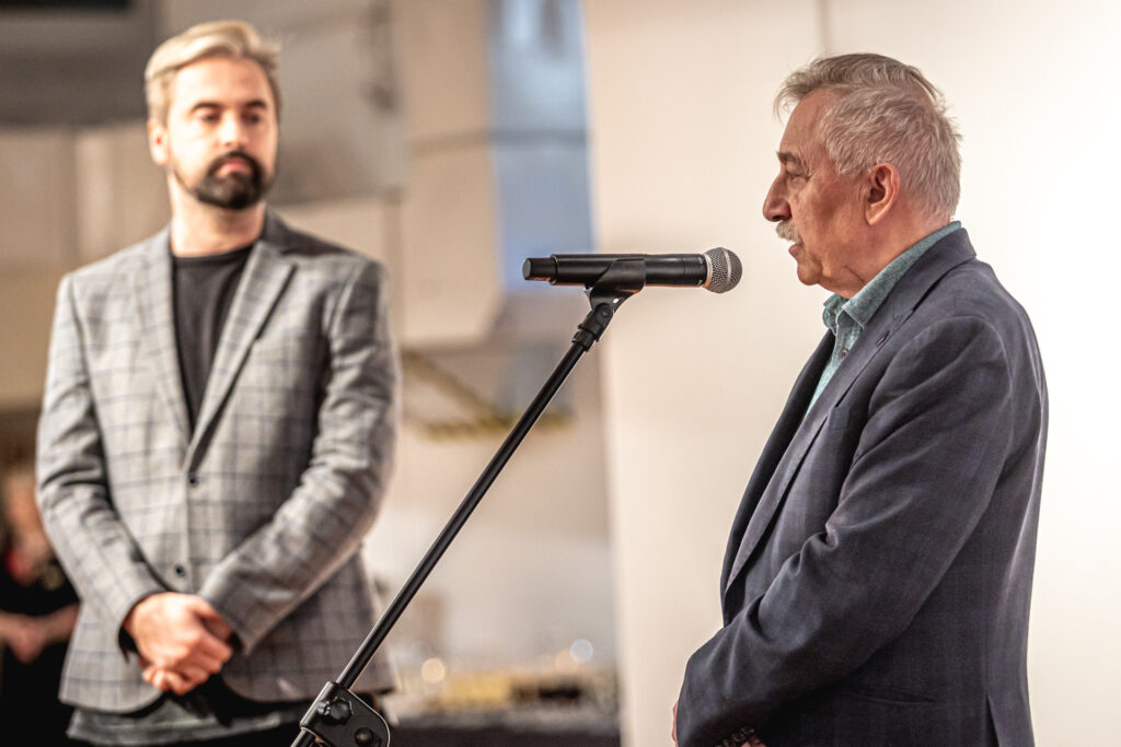 Zdjęcie. Starszy pan o siwych włosach i siwych wąsach mówi do mikrofonu. W tle wicedyrektor KCK Artur Wijata.
