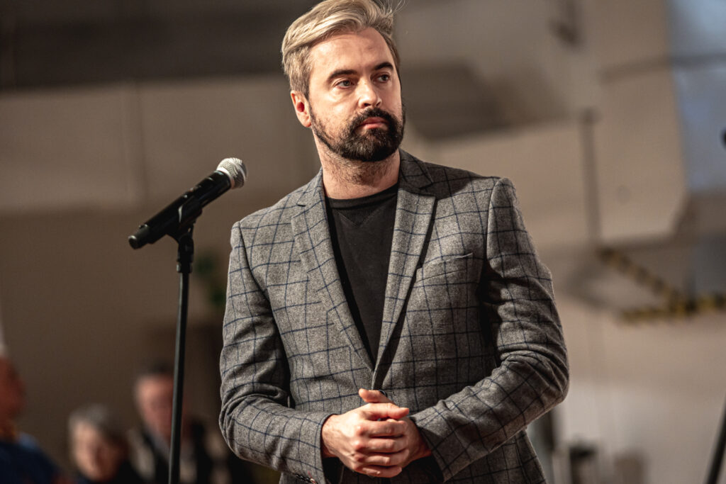 Zdjęcie. Wicedyrektor Kieleckiego Centrum Kultury. Mężczyzna ubrany w szarą marynarkę w ciemną kratę i czarny T-shirt. Ma blond włosy i ciemną brodę. Stoi przed mikrofonem. To Artur Wijata.
