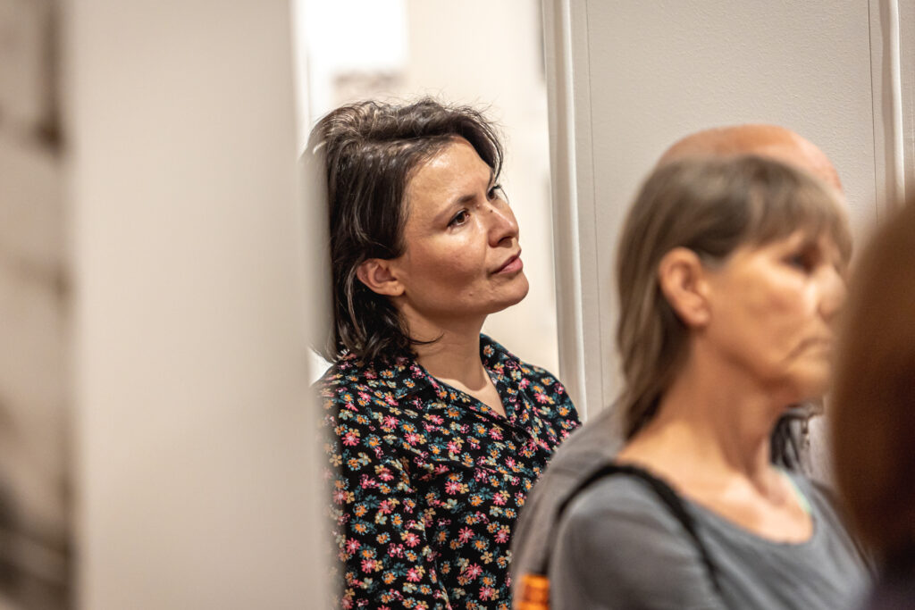 Zdjęcie. Ciemnowłosa kobieta (kierowniczka galerii Winda Daria Maroń-Ptak) patrzy przed siebie wśród innych gości wernisażu wystawy Gudzowatego.