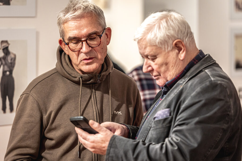 Zdjęcie. Dwóch panów w średnim wieku stoi na wystawie. Jeden z panów pokazuje drugiemu coś na telefonie komórkowym.