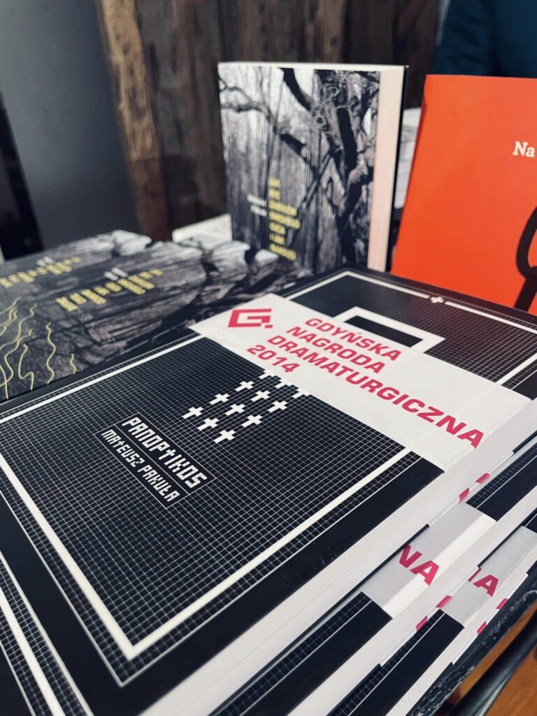 Zdjęcie. Książki Mateusza Pakuły leżące na stoliku w Galerii Winda. Na książece "Panoptikos" biała obpaska z napisem: Gdyńska Nagroda dramaturgiczna 2014