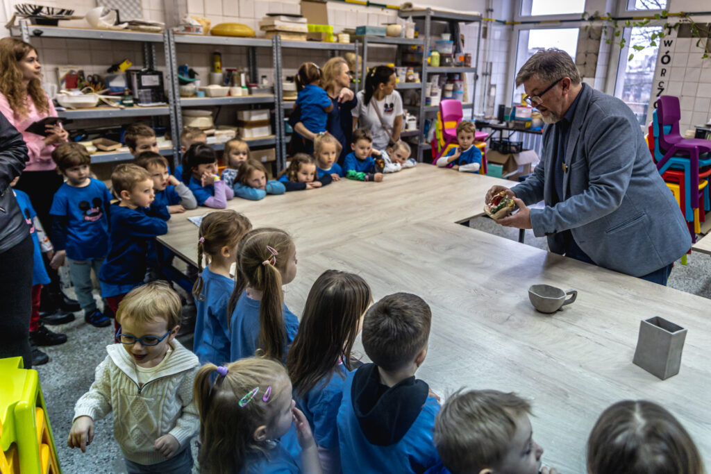 Zdjęcie. Robert Wieczorek w pracowni ceramicznej pokazuje dzieciom ceramiczną szkliwioną figurkę.