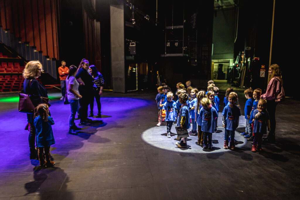 Zdjęcie. Duża Scena KCK. Duża grupa dzieci w wieku przedszkolnym stoi w grupie. Oświetla ich jeden z reflektorów.