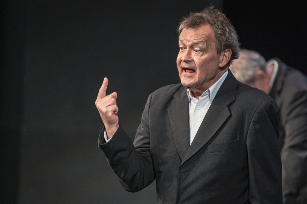 Zdjęcie. Jan Frycz Podnosi do góry wskazujący palec prawej ręki. Patrzy w kierunku palca i mówi coś z miną szalonego naukowca.
