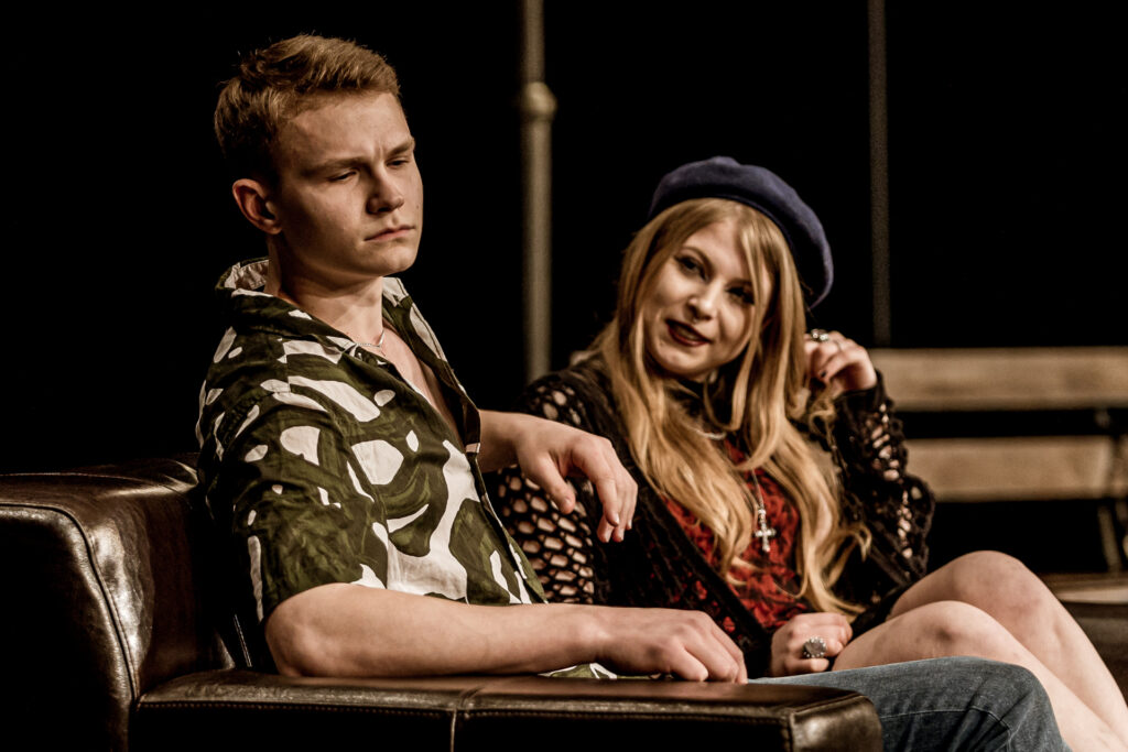 Zdjęcie. Spektakl teatru Pegaz. Dwójka młodych ludzi siedzi na kanapie. Mężczyna odwraca wzrok od kobiety z zaciętą miną. Dziewczyna w niebieskim berecie przygląda się chłopakowi z uśmiechem.