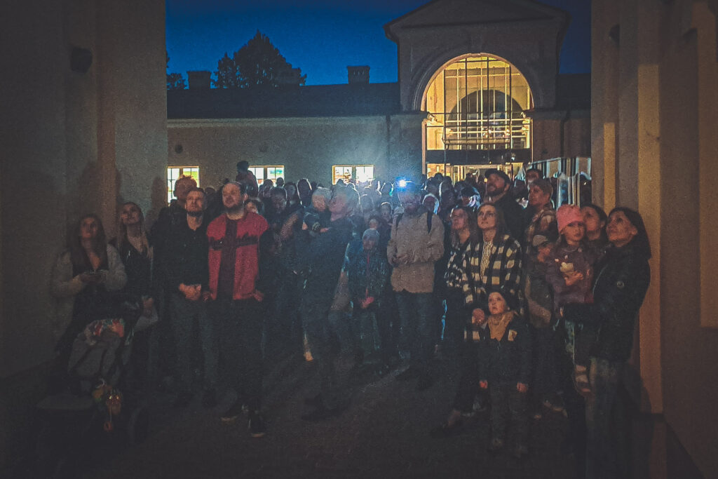 Zdjęcie. Biała Noc w Muzeum Zabawek i Zabawy. Licznie zgromadzona publiczność obserwuje występ Studia Pantomimy w oświetlonym szklanym łączniku w bramie instytucji.