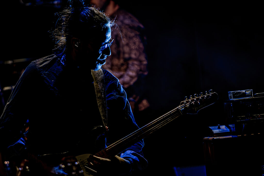 Zdjęcie. Koncert zespołu Sztywny Pal Azji. Zdjęcie. Ukryty w mroku i oświetlony jedynie niebieskim światłem sceny mężczyzna z długimi włosami spiętymi na czubku głowy w kok. Mężczyzna gra na gitarze.