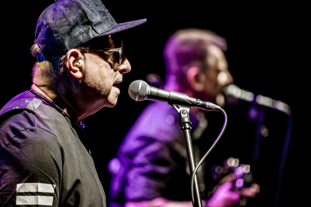 Zdjęcie. Koncert zespołu Sztywny Pal Azji. Mężczyzna w bejsbolówce śpiewa do mikrofonu. W tel kolejny muzyk śpiewa do mikrofonu.