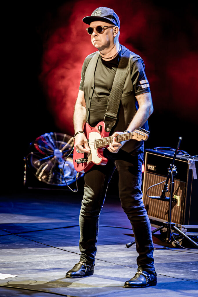 Zdjęcie. Koncert zespołu Sztywny Pal Azji. Mężczyzna w średnim wieku, ciemnych okularach z bejsbolówką na głowie gra na czerwonej gitarze elektrycznej i śpiewa do mikrofonu.