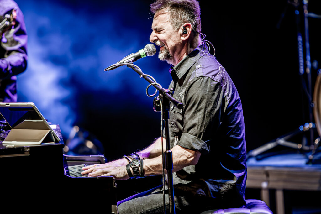 Zdjęcie. Koncert zespołu Sztywny Pal Azji. Mężczyzna z brodą siedzi przy pianinie. Śpiewa do mikrofonu.