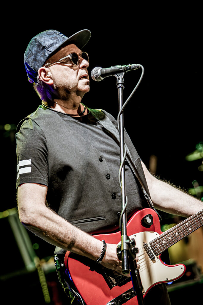 Zdjęcie. Koncert zespołu Sztywny Pal Azji. Mężczyzna w średnim wieku, ciemnych okularach z bejsbolówką na głowie gra na czerwonej gitarze elektrycznej i śpiewa do mikrofonu.