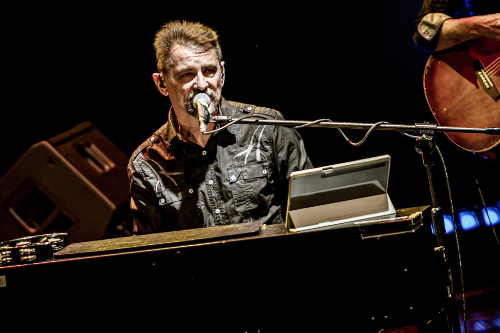 Zdjęcie. Koncert zespołu Sztywny Pal Azji. Mężczyzna z brodą siedzi przy pianinie. Śpiewa do mikrofonu.