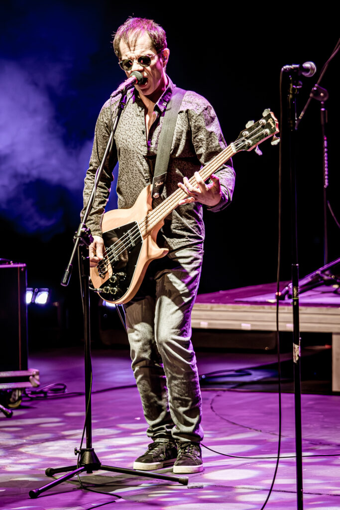 Zdjęcie. Koncert zespołu Sztywny Pal Azji. Szczupły mężczyzna w okularach przeciwsłonecznych gra na gitarze i śpiewa do mikrofonu. Oświetla go różowe światło sceniczne.