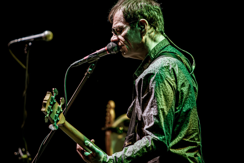 Zdjęcie. Koncert zespołu Sztywny Pal Azji. Szczupły mężczyzna w okularach przeciwsłonecznych gra na gitarze i śpiewa do mikrofonu. Oświetla go zielone światło sceniczne.