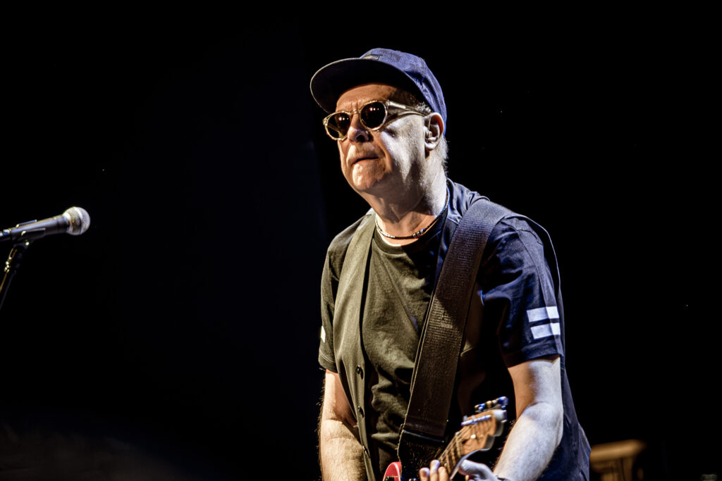 Zdjęcie. Koncert zespołu Sztywny Pal Azji. Mężczyzna w średnim wieku, ciemnych okularach z bejsbolówką na głowie gra na czerwonej gitarze elektrycznej.