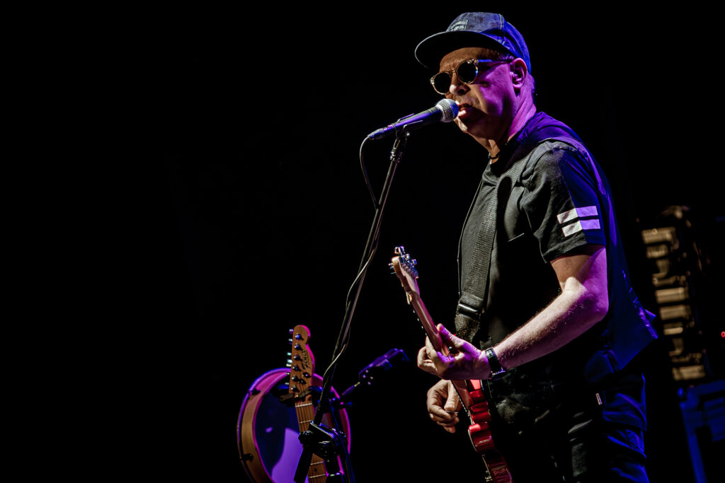 Zdjęcie. Koncert zespołu Sztywny Pal Azji. Mężczyzna w średnim wieku, ciemnych okularach z bejsbolówką na głowie gra na czerwonej gitarze elektrycznej.